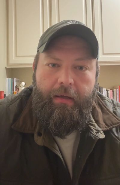 Bearded man wearing a cap and jacket in a cozy room with bookshelves.