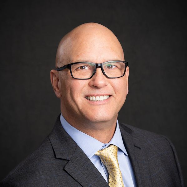 Smiling bald man wearing glasses and a suit with a yellow tie.