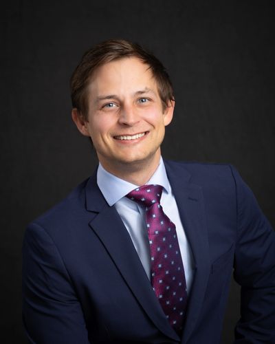 A smiling young man in a navy suit and patterned purple tie against a black background.
