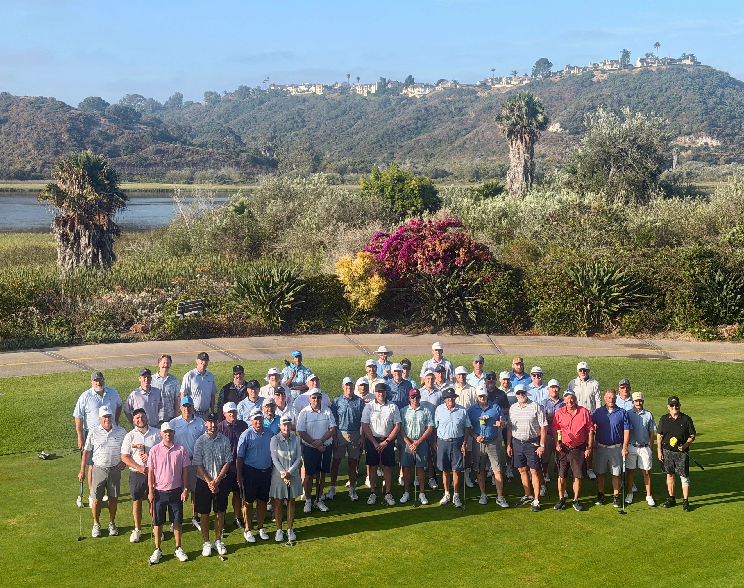 Large group of golfers posing on a green golf course with hills in the background.