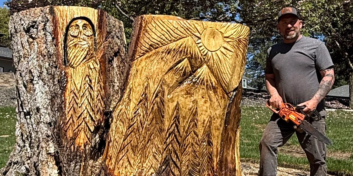 Man with chainsaw stands by detailed wood carvings on tree stumps.