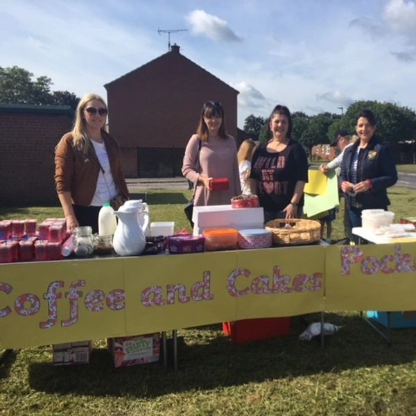 Four women standing behind a coffee and cakes stall outdoors.