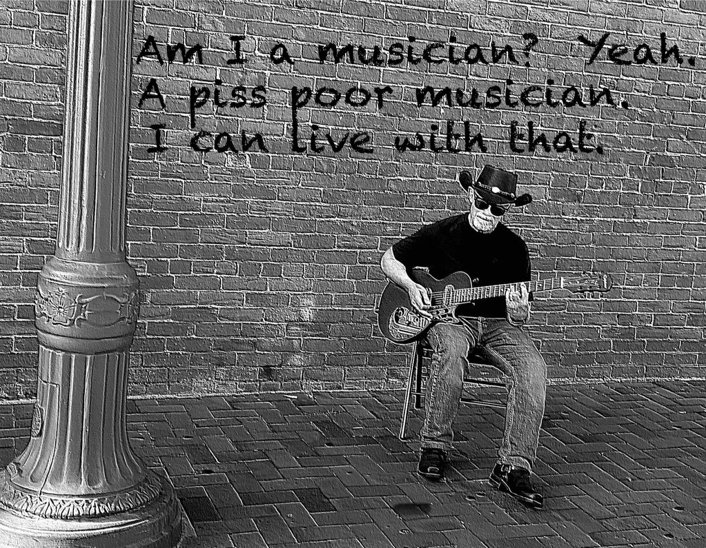 Street musician with a guitar plays in front of a brick wall with a candid message.