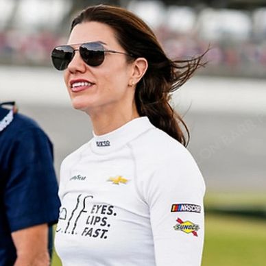 Woman in racing suit and sunglasses smiling outdoors.