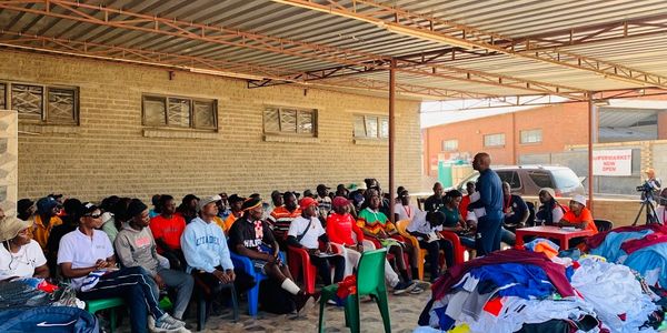 A group of people seated outdoors listening to a speaker beside piles of clothes.