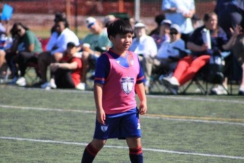 A young boy in a pink sports vest on a soccer field with spectators behind.