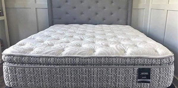 A plush, tufted mattress on a gray upholstered bed frame with a nailhead trim headboard.
