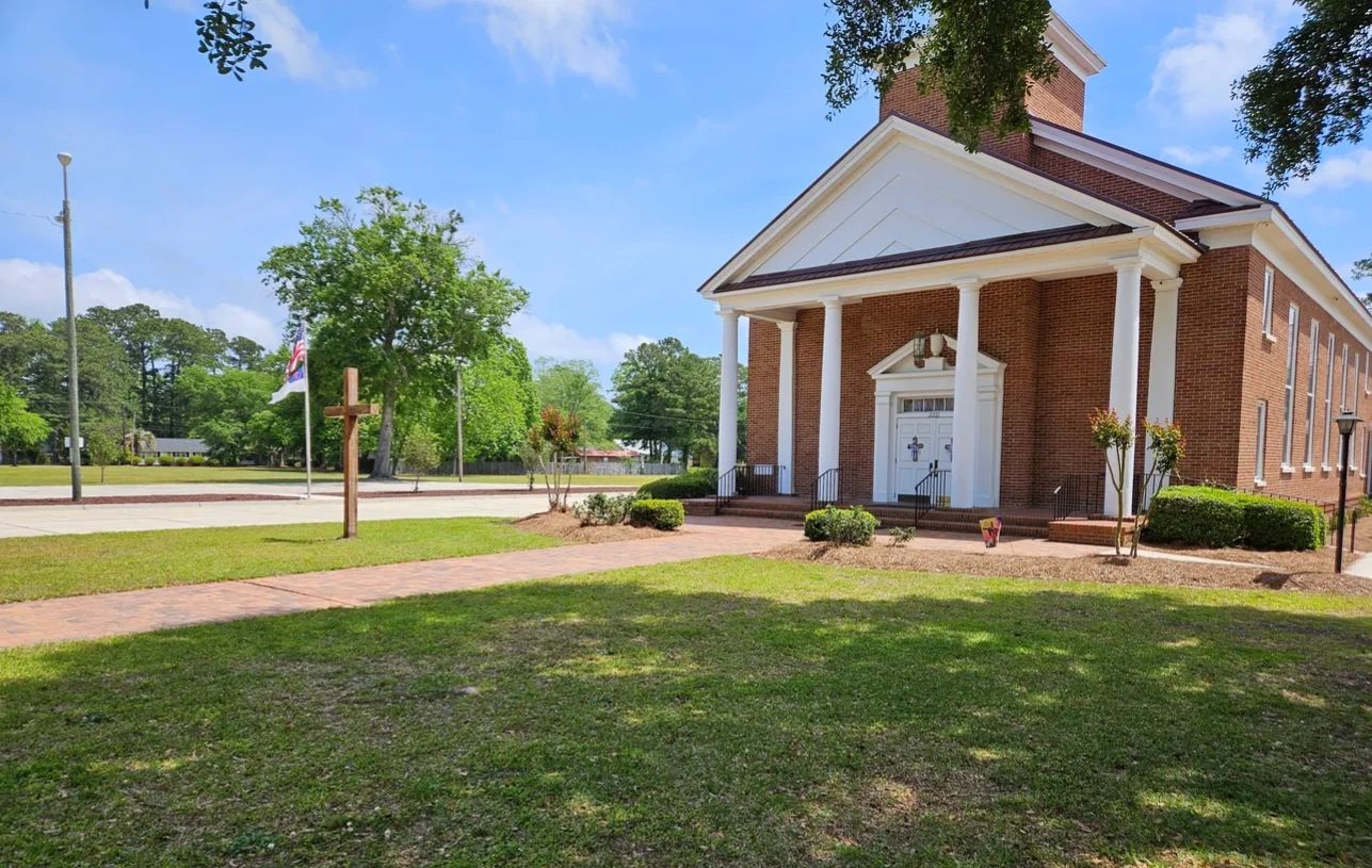 A brick church with white pillars and a wooden cross outside on a sunny day.