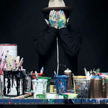 Person with paint-covered hands covering their face behind a table full of paint cans and brushes.