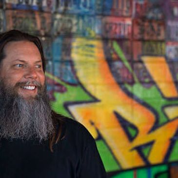 Smiling man with long beard in front of colorful graffiti wall.