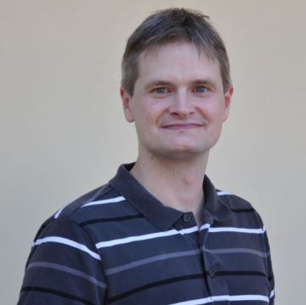 Smiling man in a black and white striped polo shirt against a plain background.