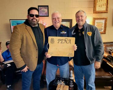 Three men posing with a wooden plaque labeled 'PEÑA' indoors.