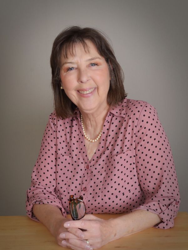 Smiling older woman in pink polka dot blouse sitting at a table.
