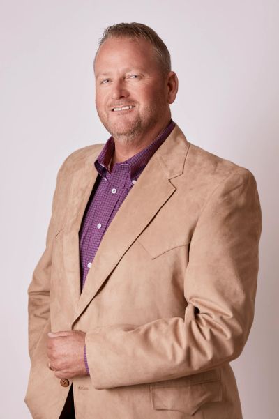 Smiling man wearing a beige jacket and purple shirt.