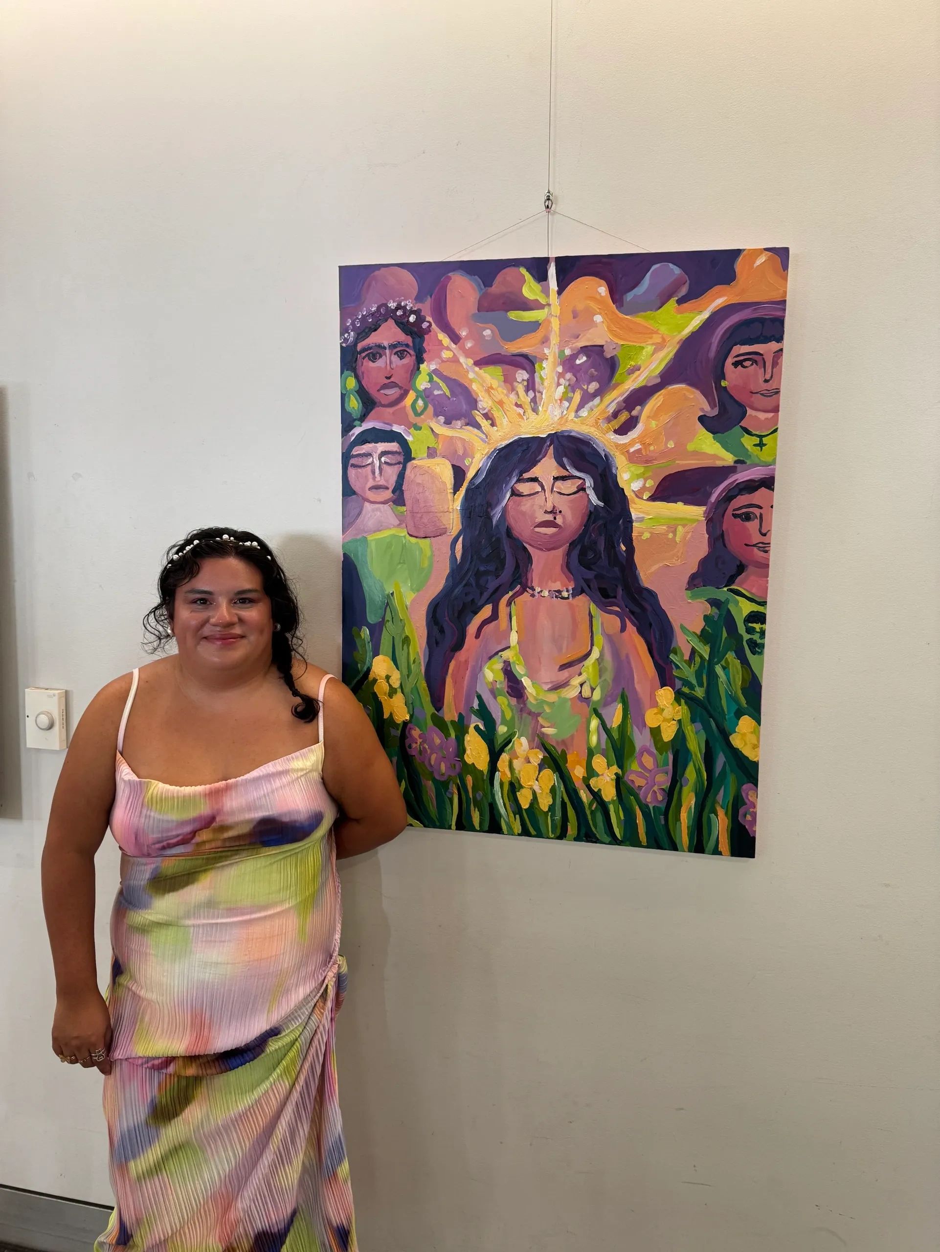 Woman in colorful dress poses next to vibrant painting of women surrounded by flowers.