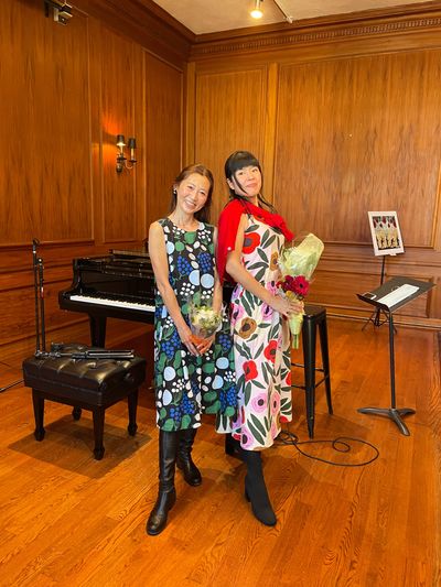 Two women in colorful dresses holding flowers in a music room.