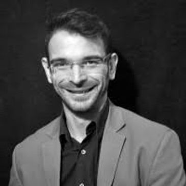 Smiling man wearing glasses and a blazer against a dark background.