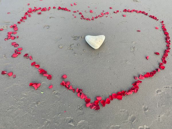 Heart shape made with red rose petals and a heart-shaped stone in the sand.