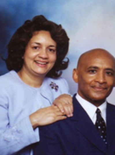 A woman and a man wearing suits, with the woman smiling and resting her hand on the man's shoulder.