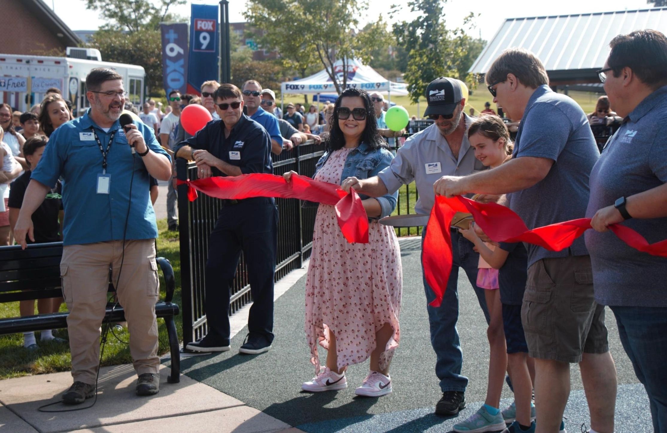 Community members cut a red ribbon at an outdoor event with a crowd and FOX 9 banners.