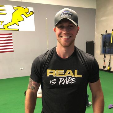 Smiling man in a gym wearing a "REAL IS RARE" shirt and a cap.