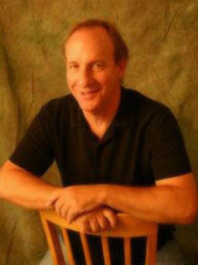 Man smiling, sitting on a wooden chair.