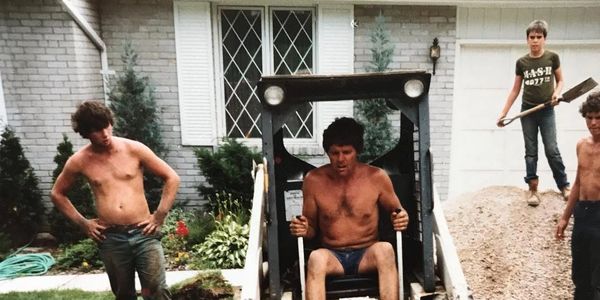 Gerry Geach Sr. operating a bobcat, while 11-year-old Gerry stands nearby holding a shovel. 