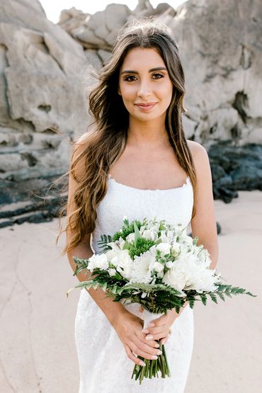 A bride in a white dress holding a bouquet on a rocky beach.