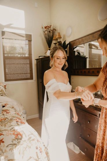 Bride in a white dress getting ready with help indoors.