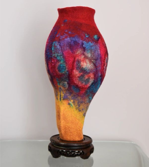 Colorful felted vase sculpture with vibrant red, purple, blue, and yellow hues on a wooden stand.