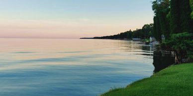 Calm lake with a grassy shore and distant trees at sunset.
