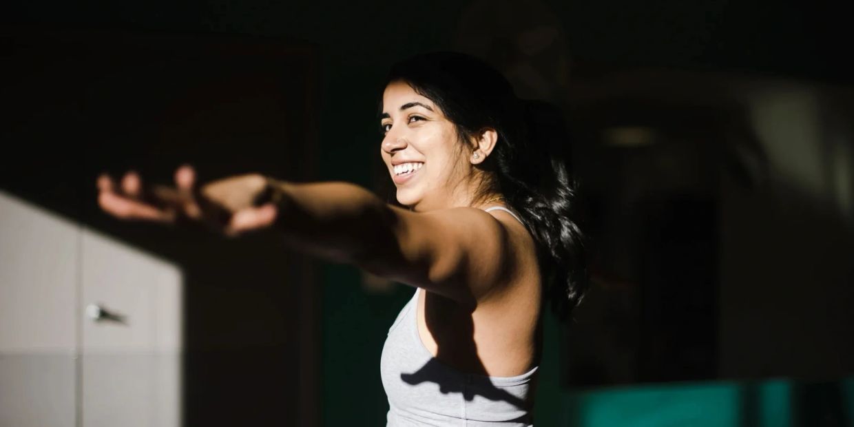 A woman smiling while stretching her arms indoors.