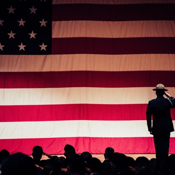 A soldier salutes in front of a large American flag with a group of people.