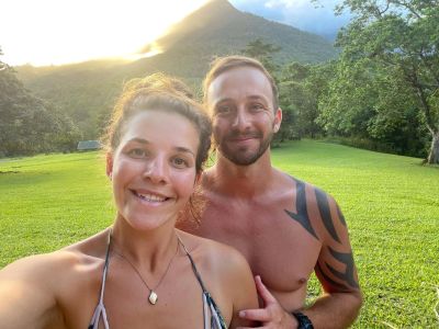 A smiling couple outdoors in a sunny, green landscape with a mountain in the background.