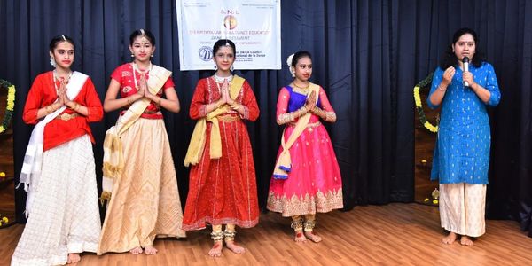 Young girls in traditional attire with a woman speaking on stage during a dance event.