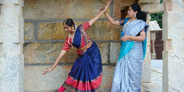 Woman teaching classical Indian dance to a student in traditional attire.