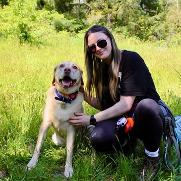 Woman with sunglasses kneeling beside a happy Labrador in a sunny grassy field.