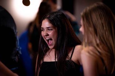 A young woman excitedly reacts with friends in a lively social setting.