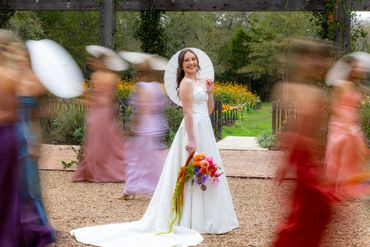 Bride in white gown holding colorful bouquet while bridesmaids move around her.