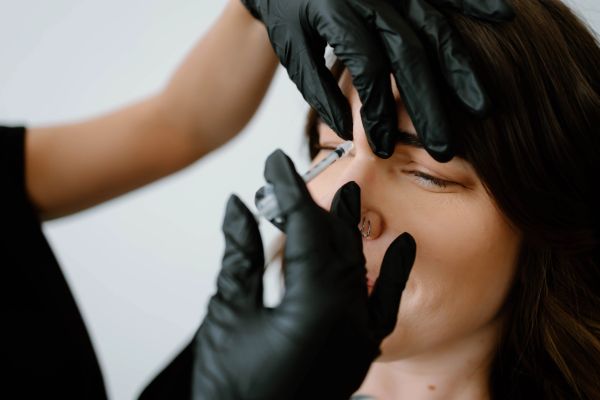 A woman receives a cosmetic injection in her nose from a professional wearing black gloves.