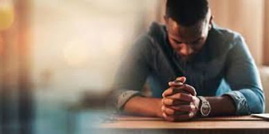 A man sits with hands clasped, head bowed in thought or prayer.