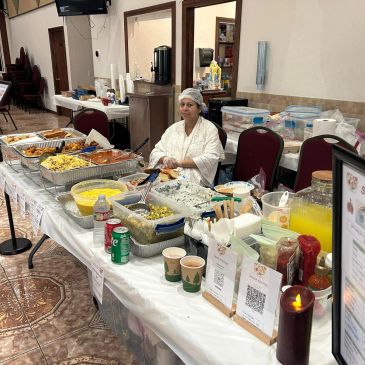 A woman in a white chef outfit sits behind a buffet table filled with various food dishes and drinks.