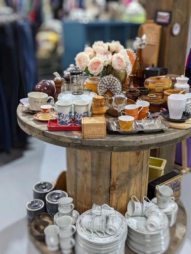 A rustic wooden table displays a variety of cups, mugs, plates, and decorative items, including flowers.