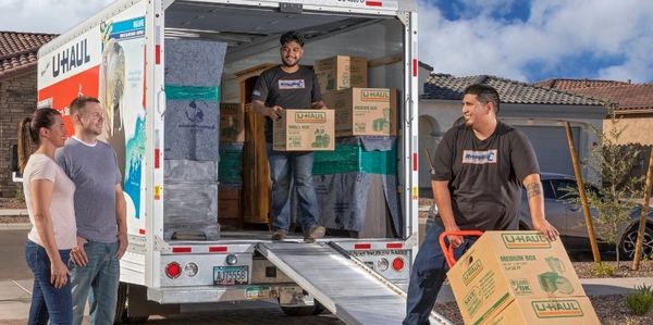 Two movers load boxes into a U-Haul truck while a couple watches.