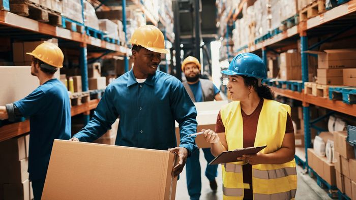 Warehouse workers collaborate, wearing safety gear and handling large boxes.
