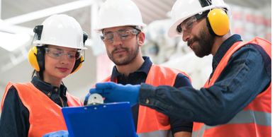 Three factory workers in safety gear reviewing a clipboard.