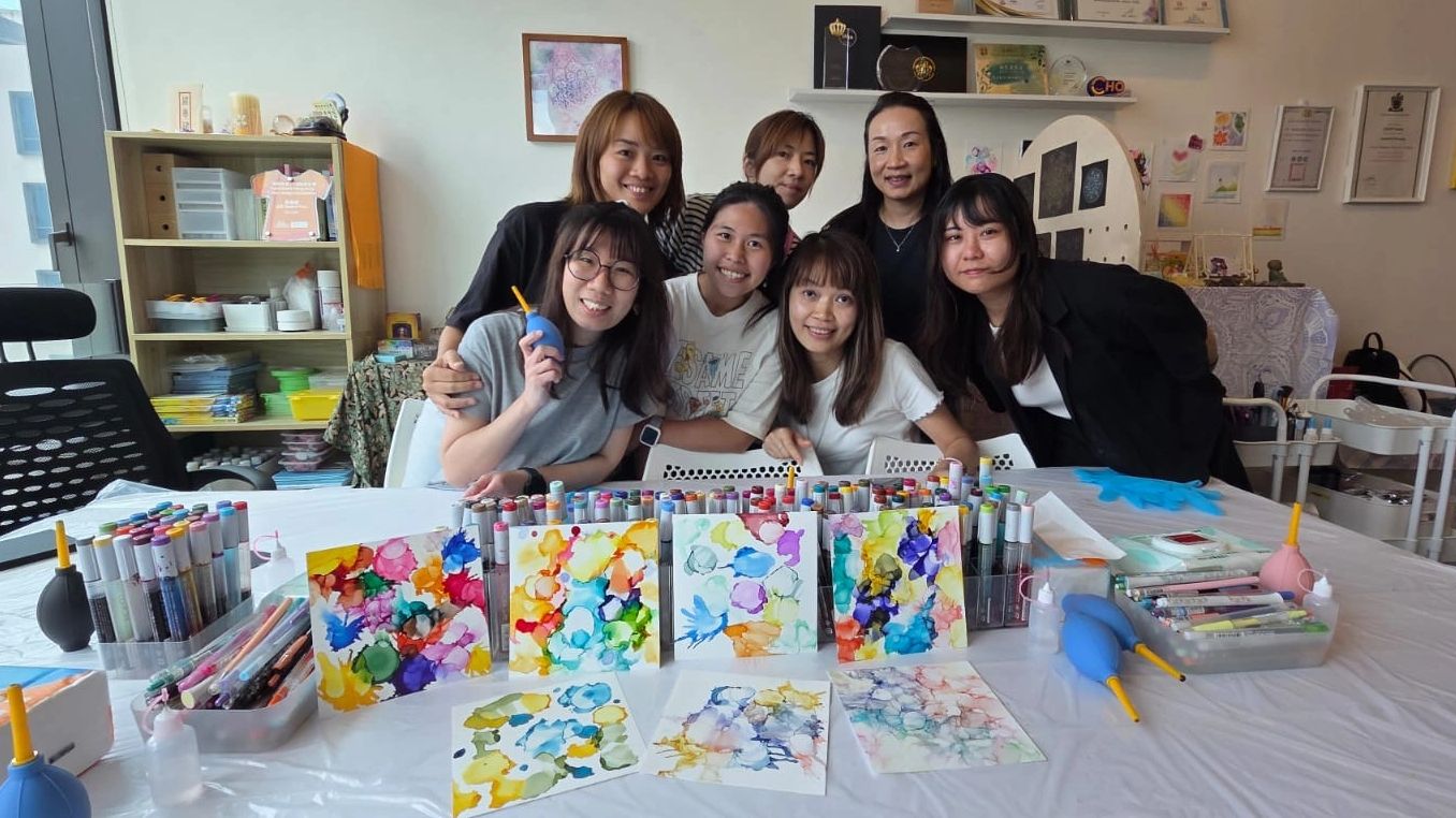 A group of seven women smiling behind colorful abstract paintings and art supplies.