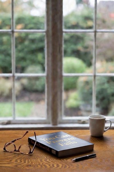Book with title, You Can Prevent Falls, resting on a table by a window with reading glasses, pen, an