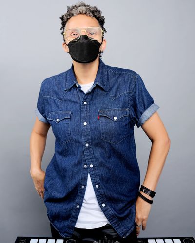 A person with light brown skin, wearing a black mask and blue denim shirt, looking at the camera. 