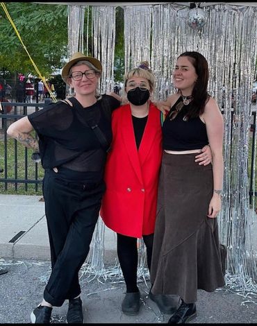 Three smiling people with arms around each other standing outside in front of silver streamers. 